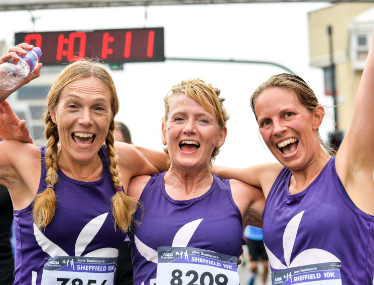 Three smiling women in matching Sheffield 10K running vests celebrate at the finish line with a race timer in the background.