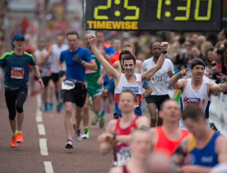 Runners approaching the finish line of a marathon, with arms raised in celebration and a large race clock displaying 2:51:30 overhead.